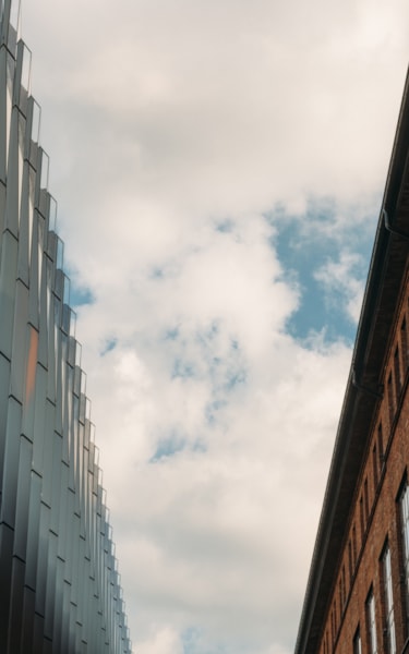 Close-up view looking up between a modern glass building and an older brick building under a partly cloudy sky.