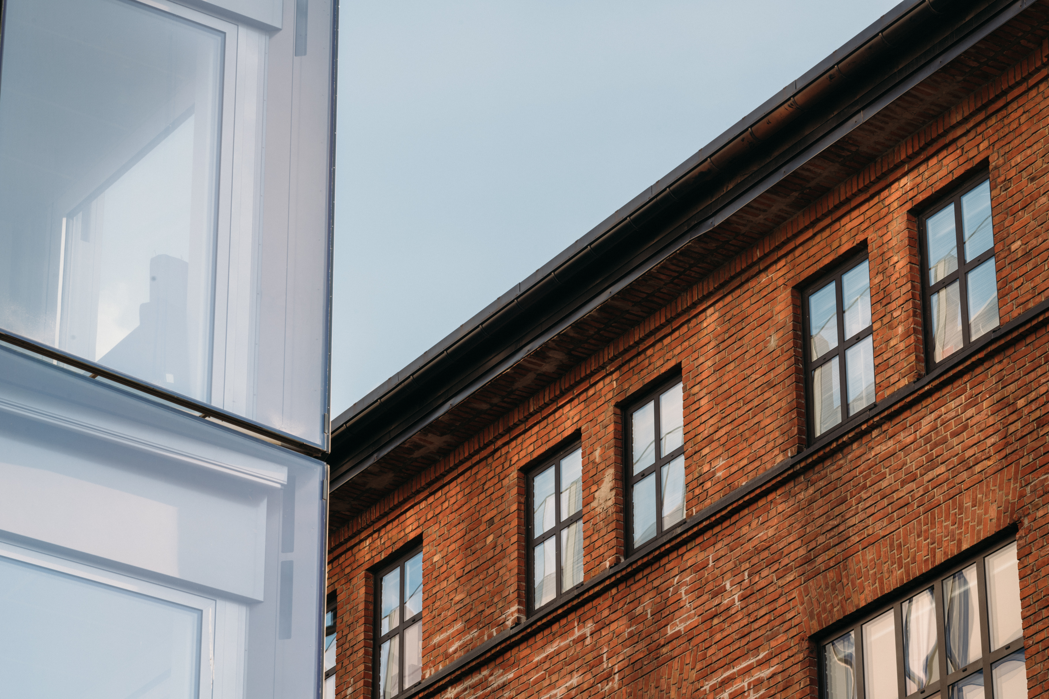 Nærbilde av en moderne glassbygning ved siden av en eldre murbygning med vinduer, med synlig himmel i bakgrunnen.