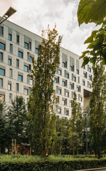 Full shot of the Diagonale building with many windows and a white facade, partly hidden by trees, a street, and a road with cars driving by.