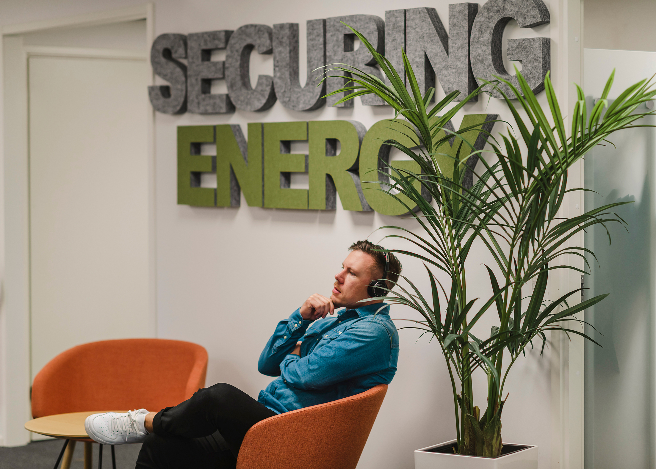 En person sitter i en oransje stol i et kontorområde. Et skilt bak dem viser teksten "SECURING ENERGY," og en høy potteplante står i nærheten.