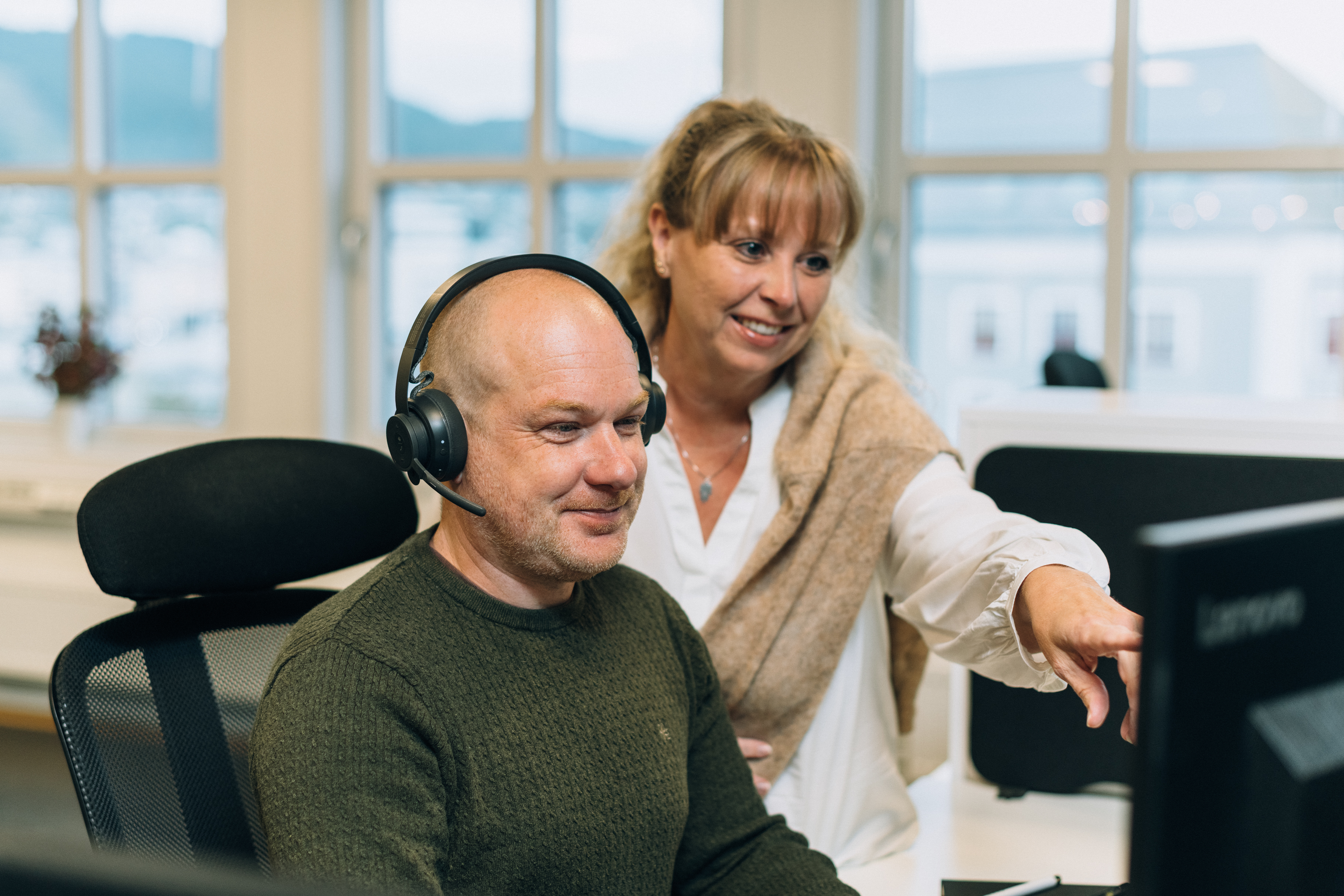 Two people in an office, one seated with headphones and the other pointing at a computer screen, with large windows in the background.