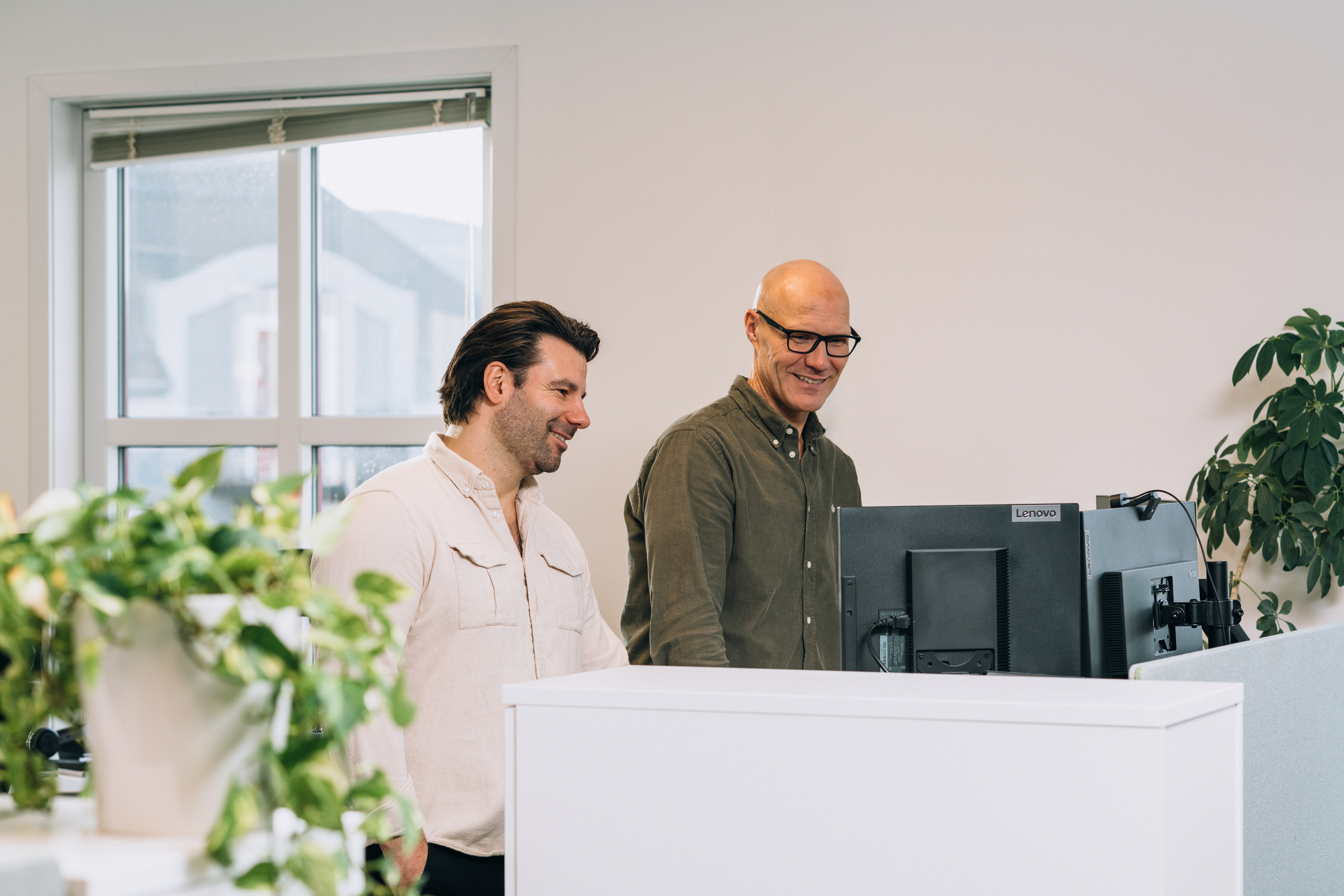 To personer står foran en Lenovo-skjerm i et kontor med planter og et vindu med delvis åpne persienner.