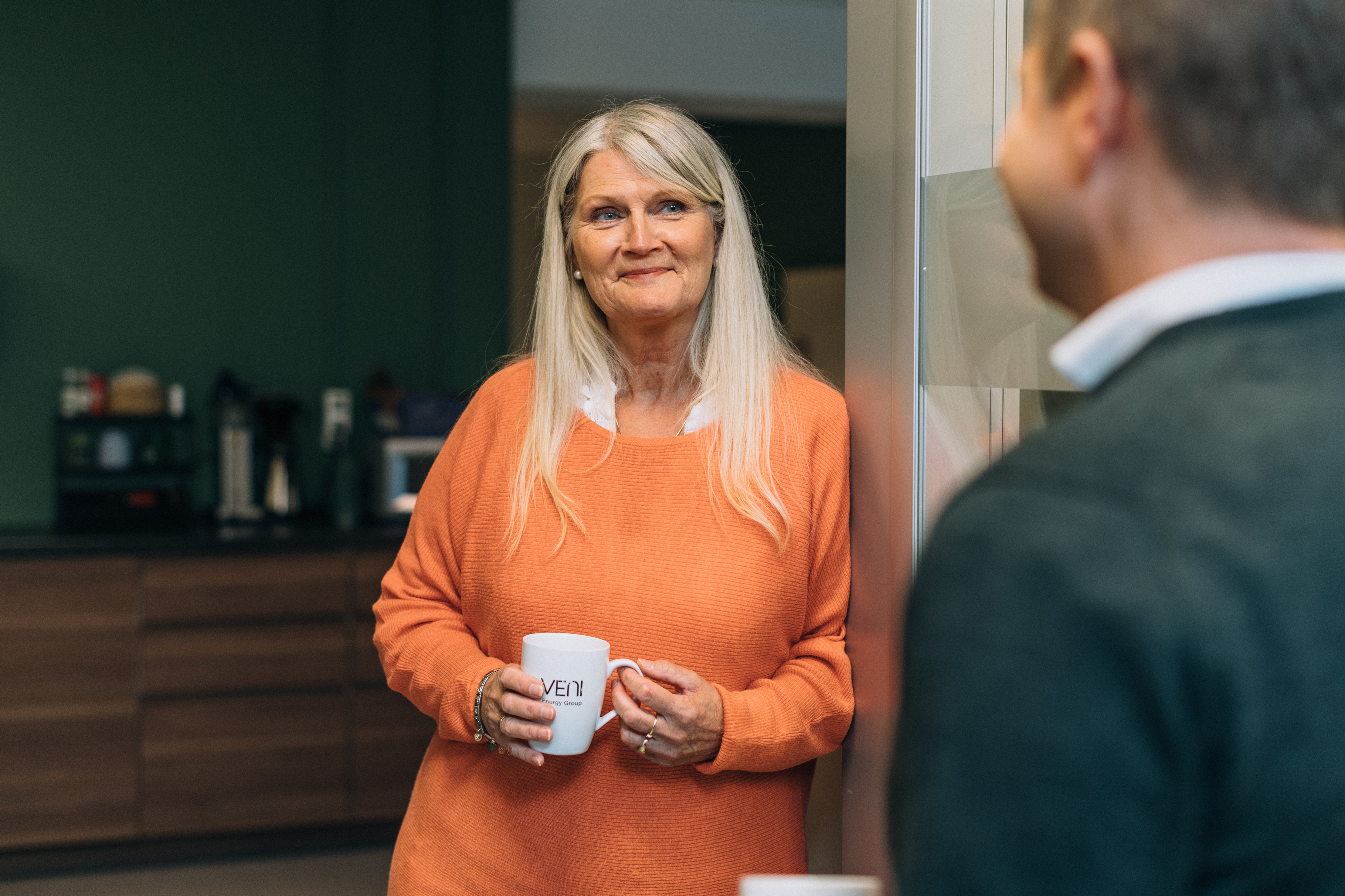 To personer står på et kjøkken. Den ene har på seg en oransje genser og holder en hvit kopp med VENI Energy Group-logoen; den andre er delvis synlig og vender mot den første.