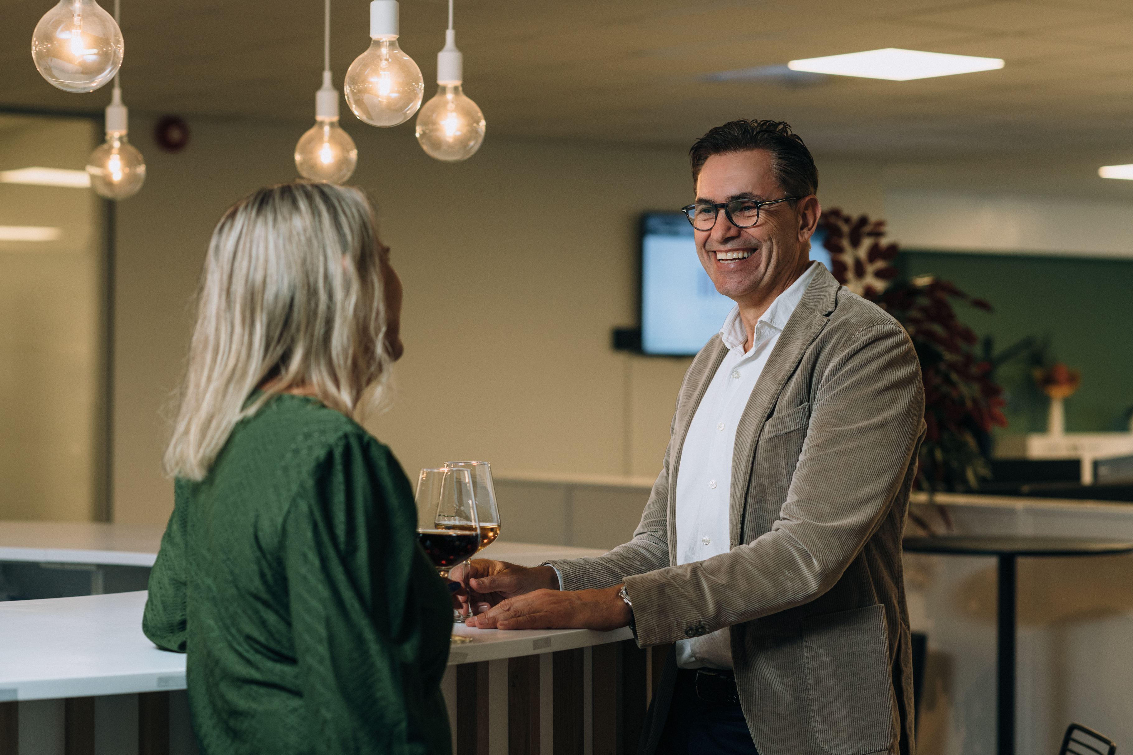 Two people stand by a bar and converse indoors, each holding a wine glass. Hanging light bulbs, a blurred screen, and plants are in the background.