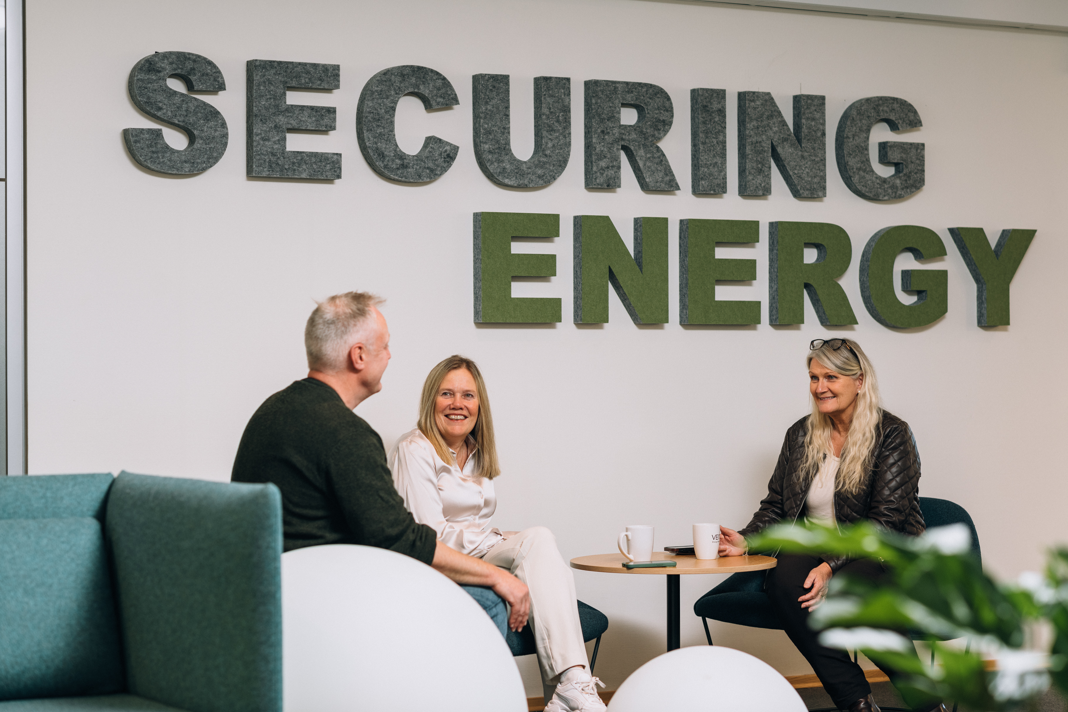 Three people sit and talk in a modern office. The wall behind them reads "SECURING ENERGY."