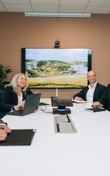 Six people in business attire sit around a conference table in a modern office, engaged in a meeting. A large screen shows a scenic landscape with hills and greenery.