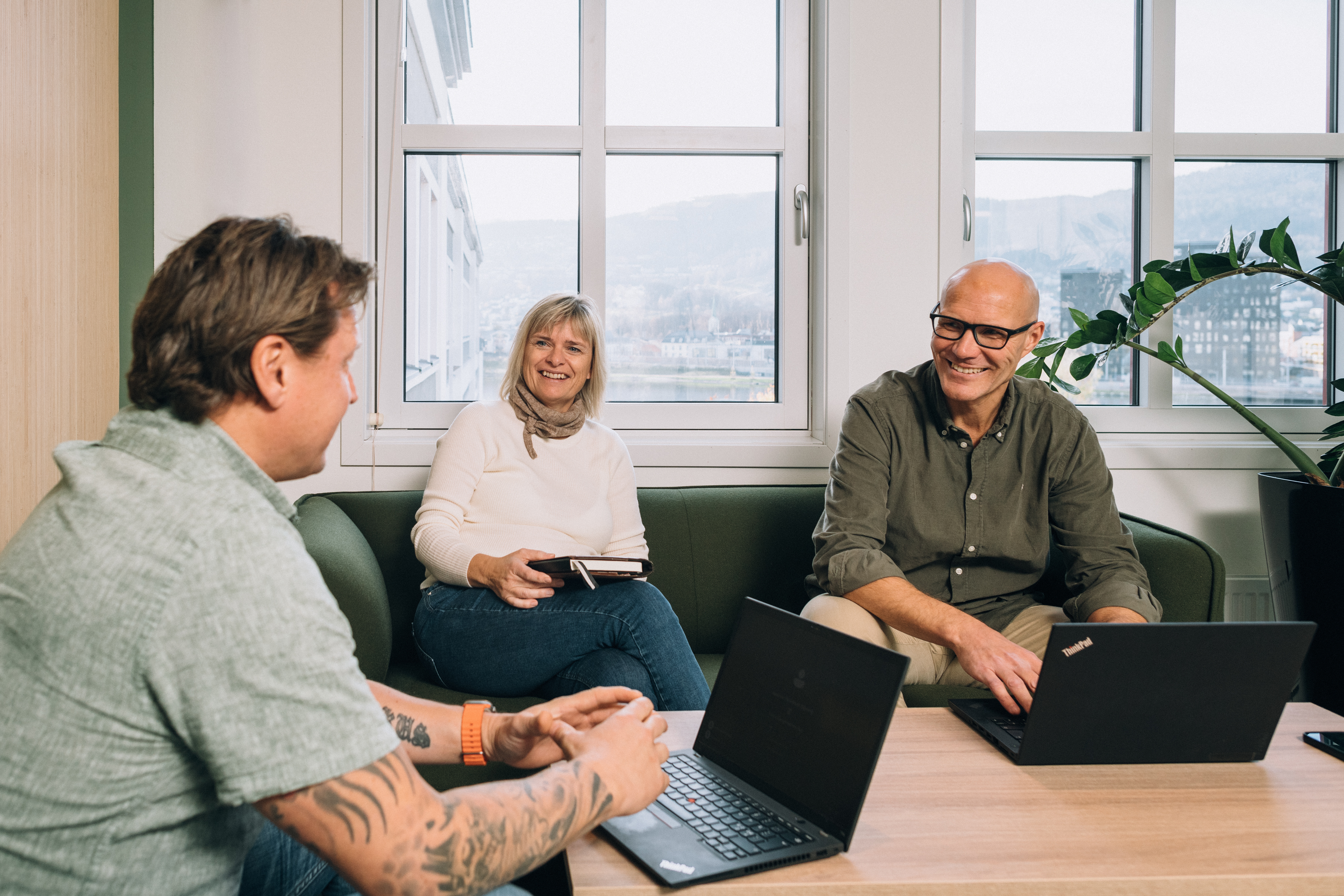 Three people sitting around a wooden table in a modern office with laptops, engaged in a discussion.
