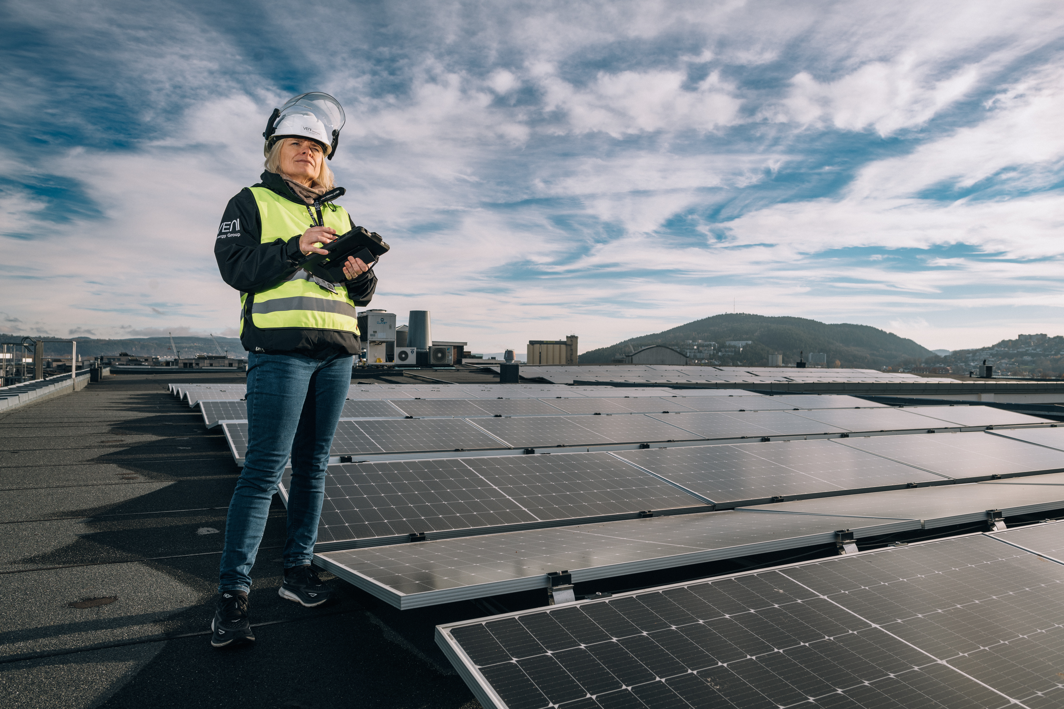 A woman wearing a safety helmet and high-visibility vest is standing on a rooftop with solar panels, holding a tablet.