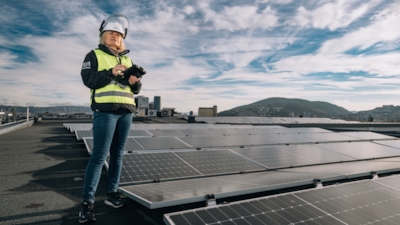 A woman wearing a safety helmet and high-visibility vest is standing on a rooftop with solar panels, holding a tablet.