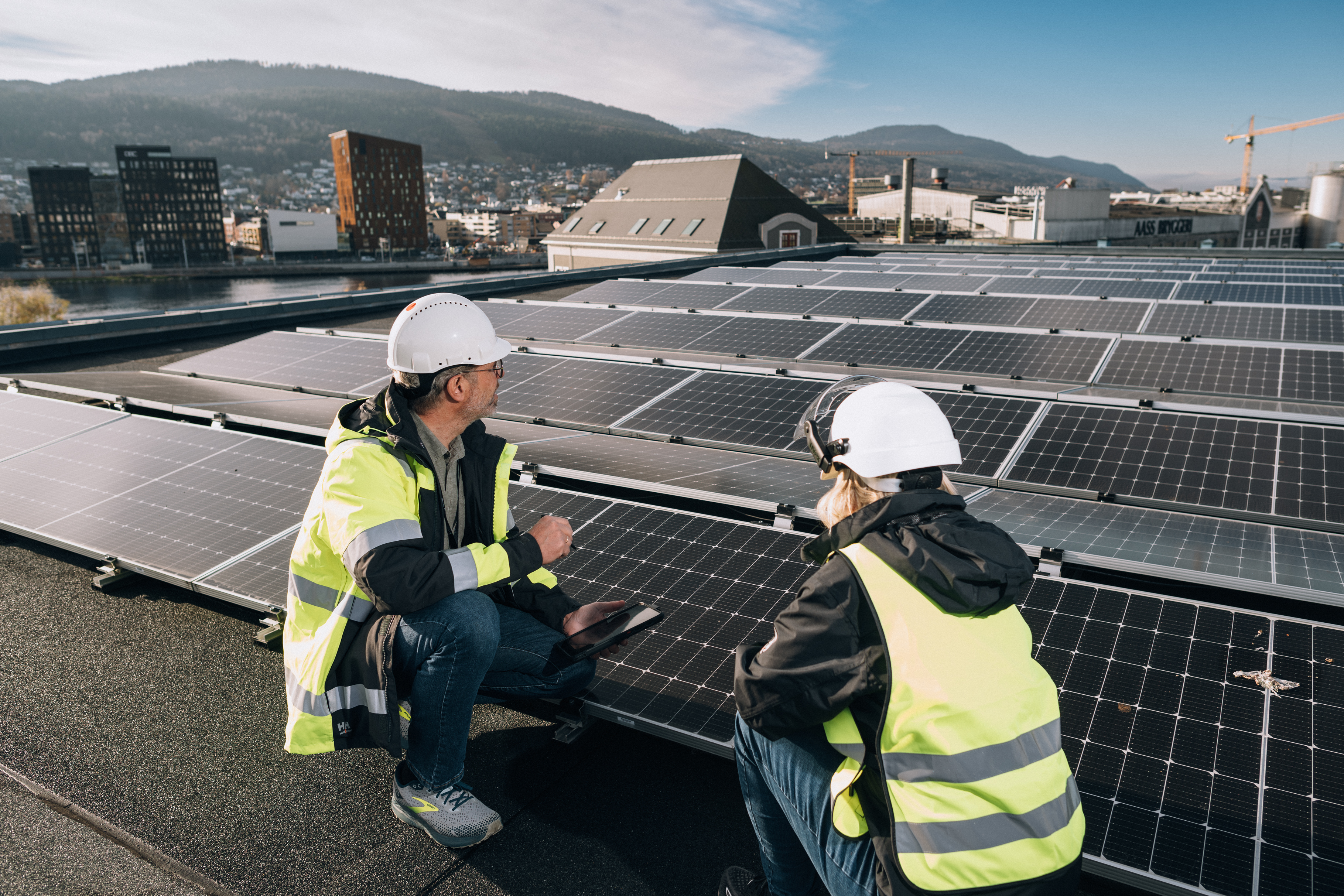 A man and a woman, both wearing safety helmets and high-visibility jackets, are kneeling on a rooftop next to solar panels. The man is holding a tablet.