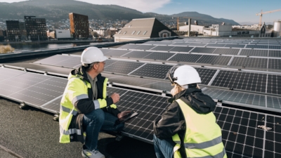 A man and a woman, both wearing safety helmets and high-visibility jackets, are kneeling on a rooftop next to solar panels. The man is holding a tablet.