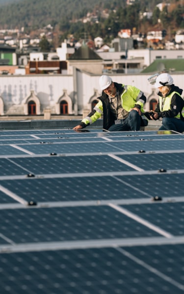 A man and a woman in safety gear inspecting solar panels on a rooftop, with buildings and hills in the background.