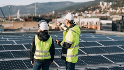 A man and a woman in safety gear standing on a rooftop with solar panels, pointing toward the cityscape with mountains in the background.