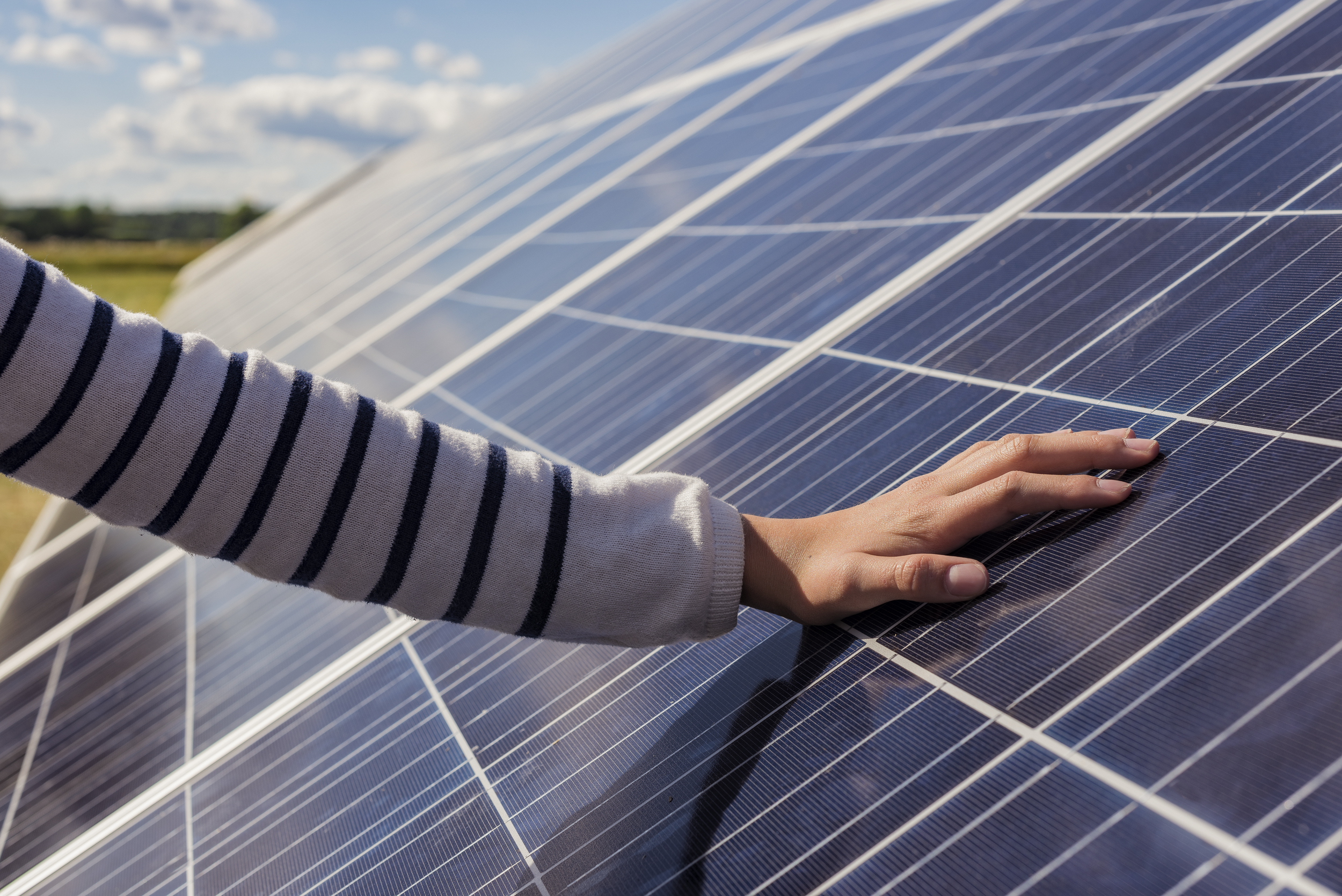 Close-up of an arm with a long-sleeved striped shirt reaching out to rest a hand on a solar panel under a partly cloudy sky.
