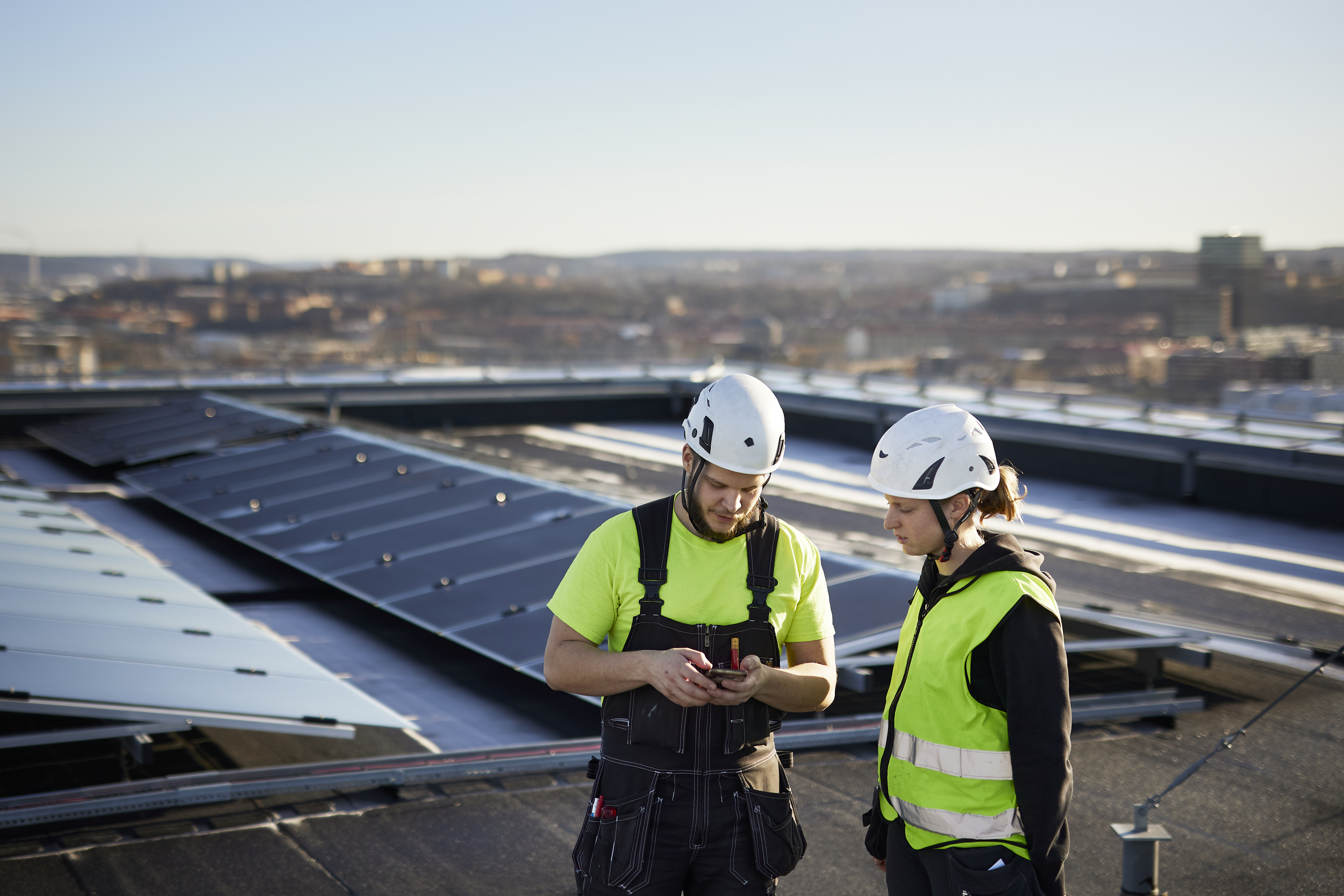 Two people in safety gear looking at a handheld device on a rooftop with solar panels and a cityscape in the background.